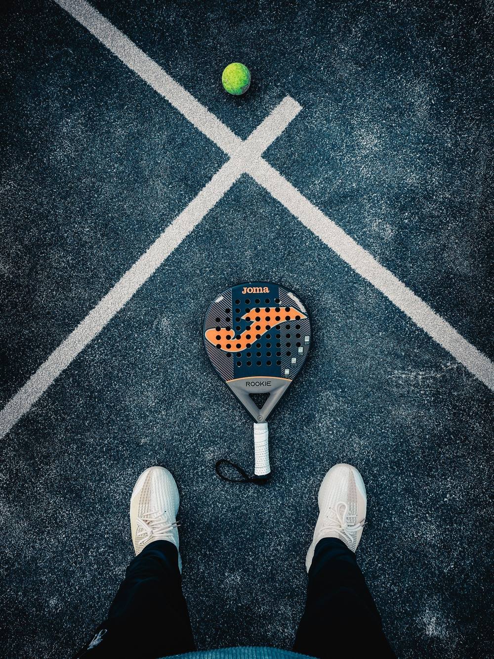 Vue de dessus d'un joueur de padel : chaussures blanches, raquette et balle posées sur le court