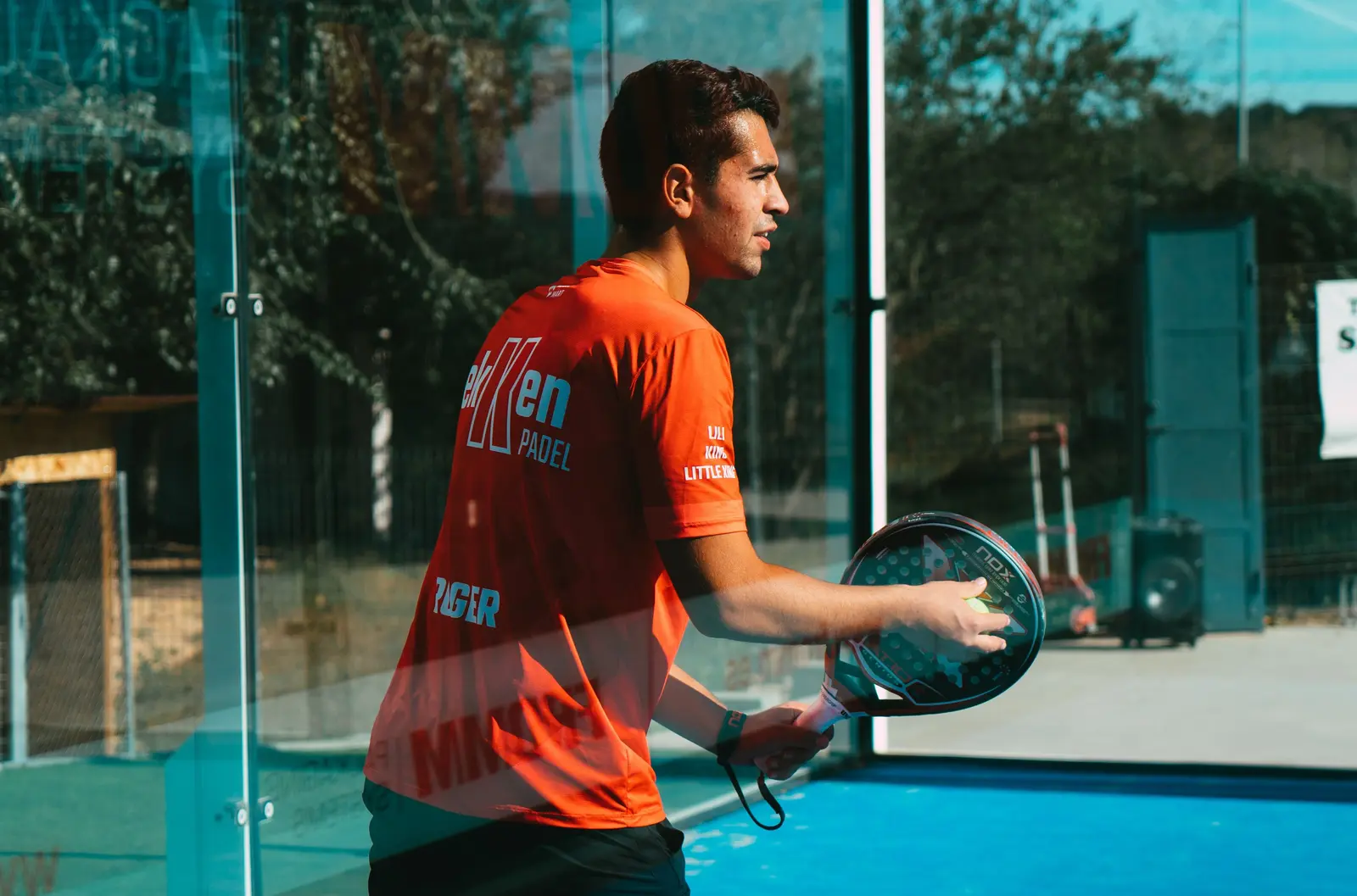 Joueur de padel en maillot technique orange, raquette en main sur un court bleu
